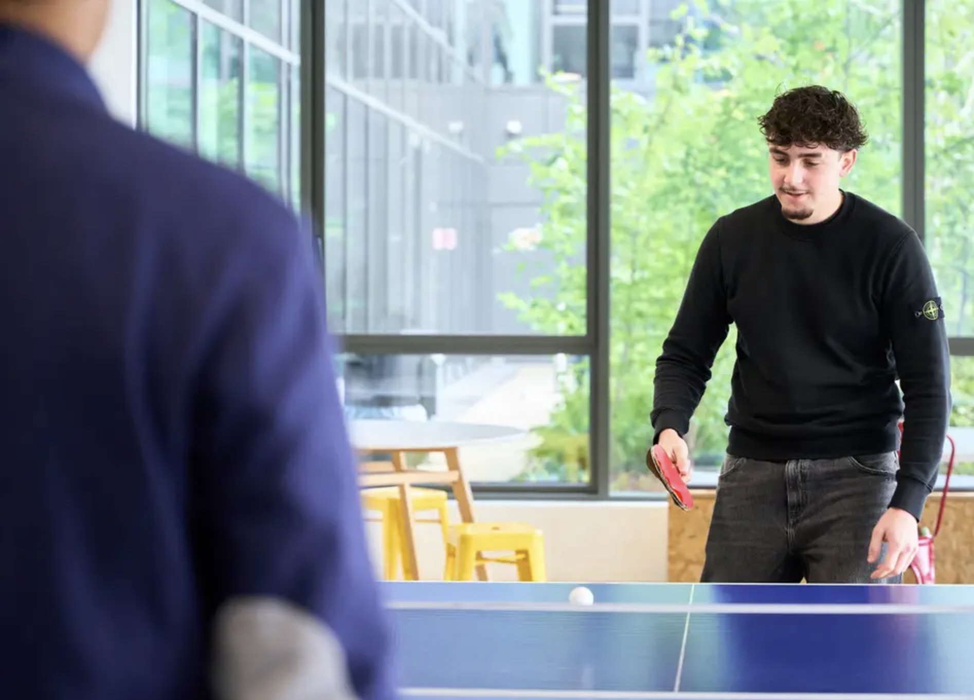 Étudiant jouant au ping-pong devant les baies vitrées du campus